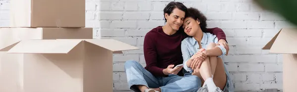 Happy multiethnic couple hugging while sitting near carton boxes, banner — Stock Photo