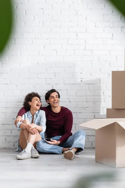 Feliz pareja multiétnica sentada en el suelo cerca de cajas de cartón - foto de stock