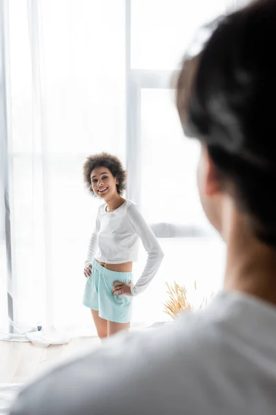 Happy african american woman standing with hands on hips while looking at blurred boyfriend — Stock Photo