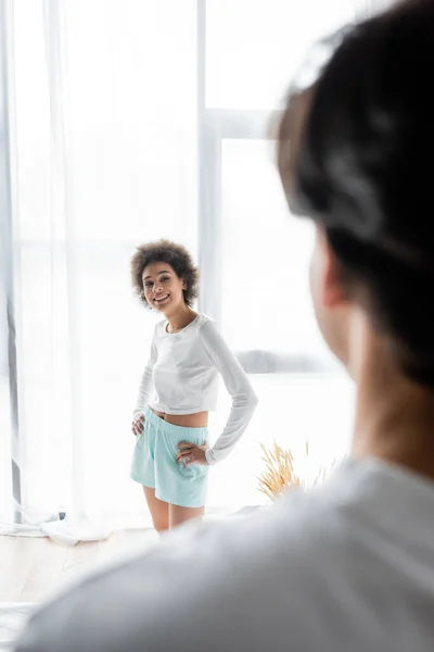 Feliz mulher afro-americana de pé com as mãos nos quadris enquanto olha para o namorado borrado — Fotografia de Stock