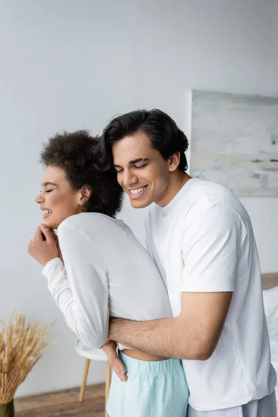 Cheerful multiethnic couple laughing and hugging in bedroom — Stock Photo