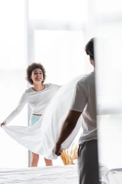 Cheerful african american woman arranging bed sheet with boyfriend — Stock Photo