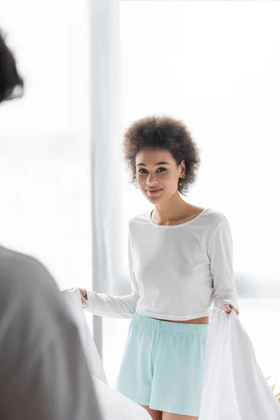 Souriant afro-américain femme organiser drap de lit avec petit ami flou — Photo de stock