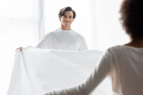Happy man arranging bed sheet with african american girlfriend — Stock Photo