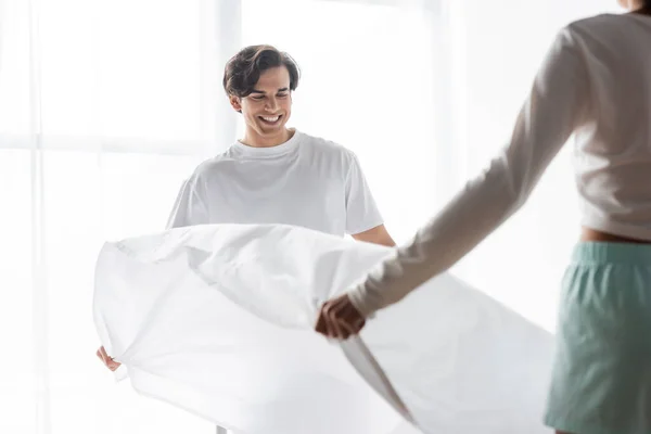 Happy man arranging bed sheet with african american girlfriend — Stock Photo