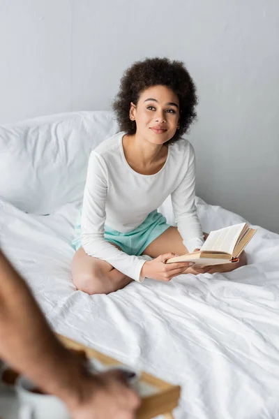 Allegra donna afroamericana seduta sul letto con libro vicino all'uomo sfocato — Foto stock