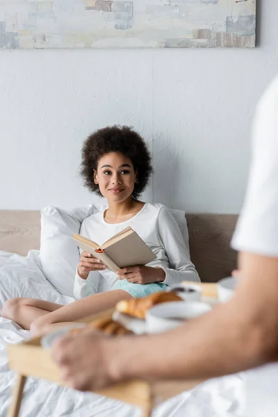 Heureux afro-américaine femme assise sur le lit avec livre près homme flou tenant plateau de petit déjeuner — Photo de stock