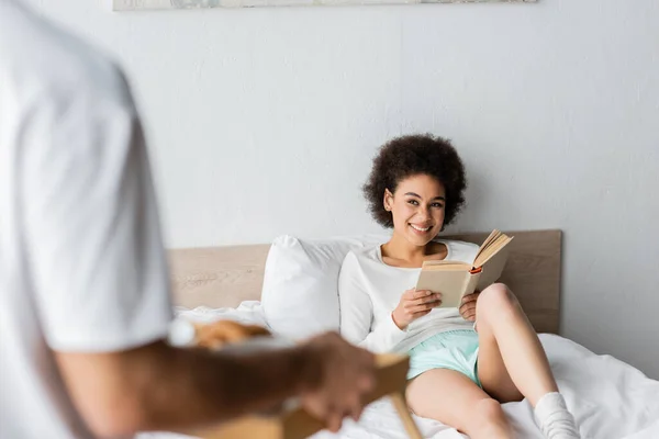 Alegre africano americano mujer sentado en cama con libro cerca borrosa hombre con desayuno bandeja - foto de stock