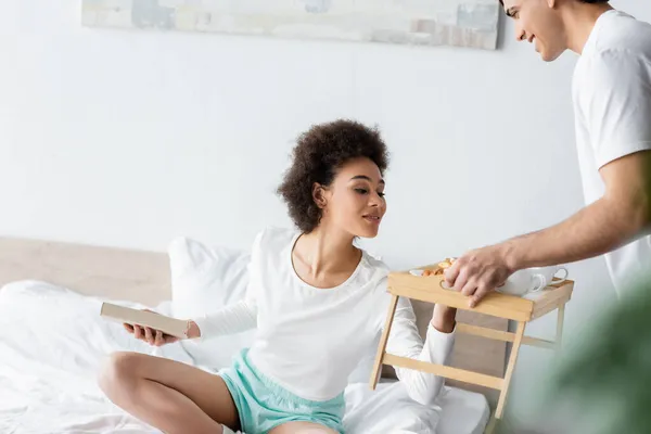 Frisé afro-américaine femme regardant plateau avec petit déjeuner dans les mains de l'homme souriant — Photo de stock