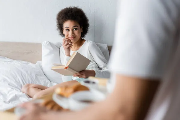 Ricci donna africana americana con libro sorridente mentre guarda l'uomo sfocato che tiene in mano il vassoio della colazione — Foto stock