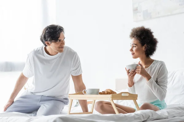 Alegre interracial casal olhando para o outro e tomando café da manhã na cama — Fotografia de Stock