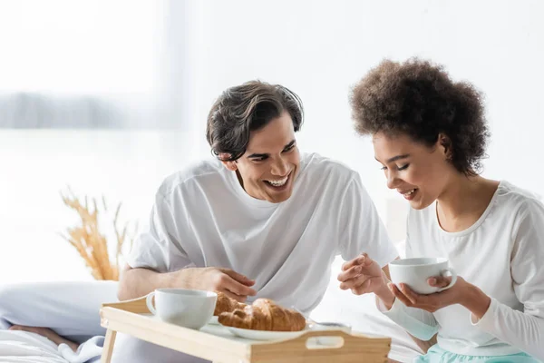 Joyeux couple interracial souriant tout en prenant le petit déjeuner au lit — Photo de stock