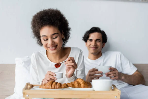 Cheerful african american woman holding spoon with jam near croissants and blurred boyfriend with cup — Stock Photo