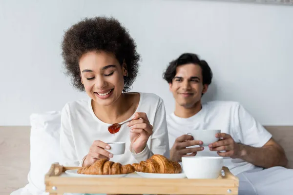 Mulher americana africana alegre segurando colher com geléia perto de croissants e namorado borrado com copo — Fotografia de Stock