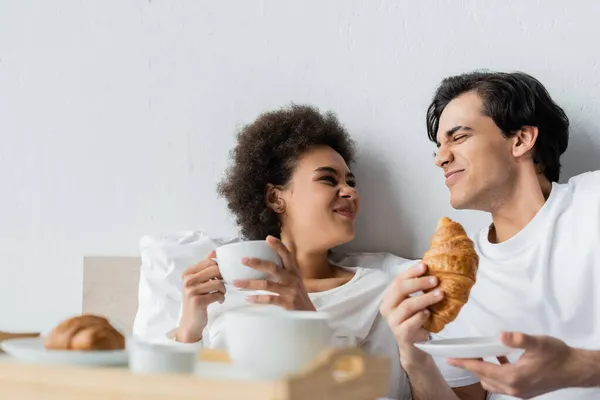 Funny interracial couple smiling and wrinkling noses while having breakfast in bed — Stock Photo