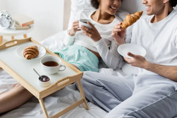 Vue recadrée de joyeux couple interracial prenant le petit déjeuner au lit — Photo de stock