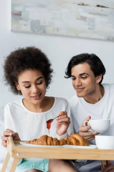 Donna afro-americana sorridente che tiene cucchiaio con marmellata vicino al fidanzato con tazza mentre fa colazione a letto — Foto stock