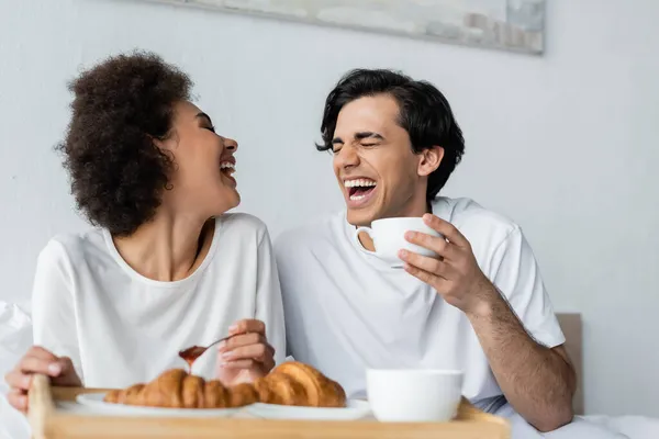 Cheerful african american woman holding spoon with jam near boyfriend with cup while having breakfast in bed — Stock Photo