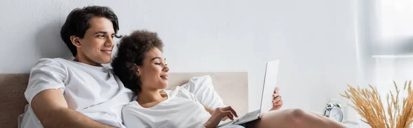 Happy african american freelancer using laptop near boyfriend in bed, banner — Stock Photo