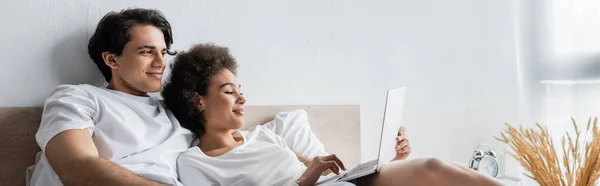 Feliz afroamericano freelancer utilizando portátil cerca de novio en la cama, bandera - foto de stock