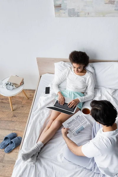 Vista superior del freelancer afroamericano usando el ordenador portátil cerca del novio leyendo el periódico de la vida de viaje en la cama - foto de stock