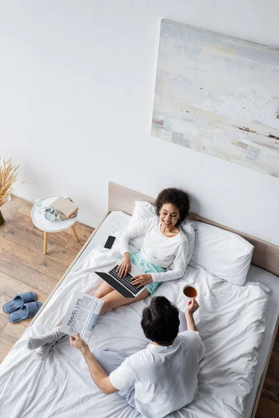 Vue du haut de heureux afro-américain pigiste en utilisant un ordinateur portable près du petit ami tenant le journal de la vie de voyage dans le lit — Photo de stock