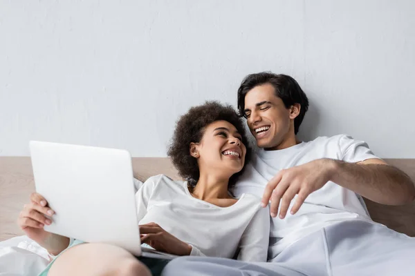 Felice freelance africano americano utilizzando laptop vicino fidanzato allegro a letto — Foto stock