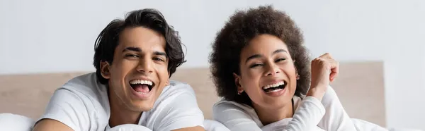 Heureux couple multiethnique rire tout en étant couché dans le lit, bannière — Photo de stock