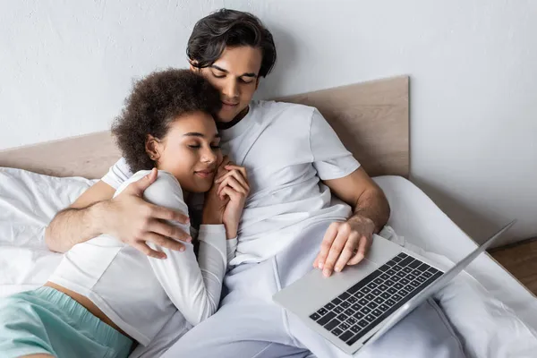 Vista de ángulo alto de la mujer afroamericana durmiendo en el pecho del novio con el ordenador portátil - foto de stock