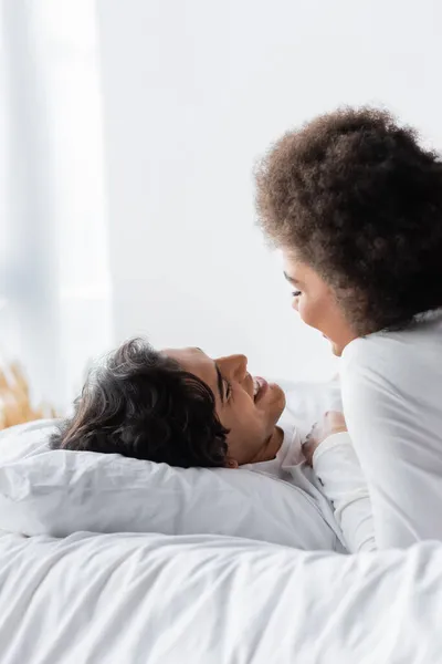 Pareja interracial positiva sonriendo mientras se miran en el dormitorio - foto de stock