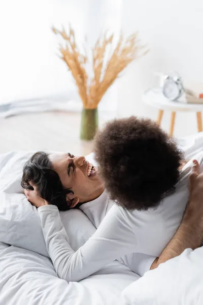 Encaracolado afro-americano mulher acariciando o cabelo do homem alegre na cama — Fotografia de Stock