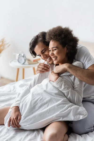 Fröhlicher Mann umarmt zufrieden afrikanische Amerikanerin auf dem Bett — Stockfoto