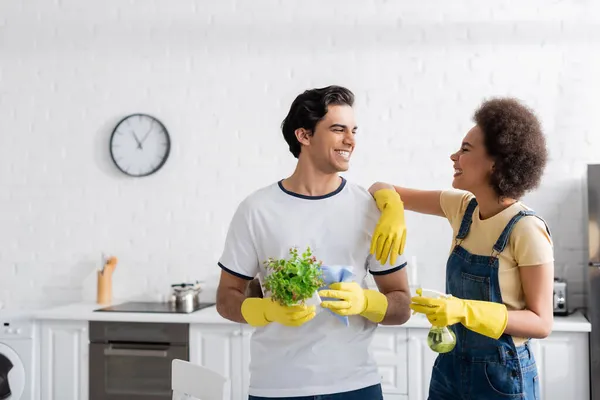 Homme heureux dans des gants en caoutchouc tenant chiffon et plante verte près de femme afro-américaine avec vaporisateur — Photo de stock
