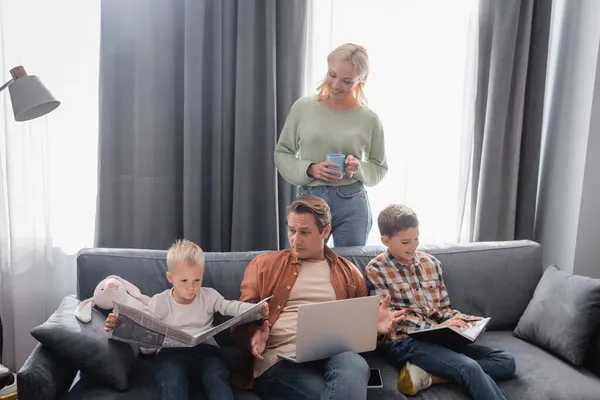 Mujer con taza de té de pie cerca de marido con portátil e hijos con cuadernos y periódico en el sofá - foto de stock