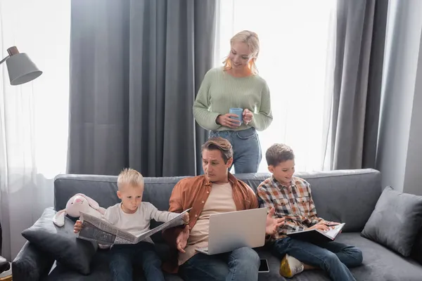Woman with cup of tea standing near husband with laptop and sons with notebooks and newspaper on sofa — Stock Photo