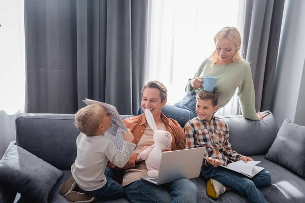 Мужчина с ноутбуком и игрушечным кроликом улыбается рядом с детьми и женой с чашкой чая в гостиной — стоковое фото