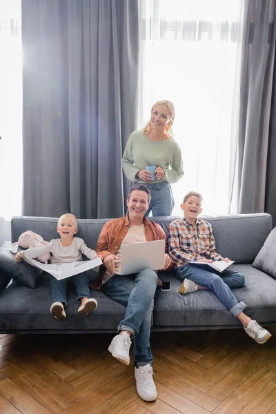 Glückliche Frau mit Tee, Mann mit Laptop und Jungen mit Zeitung und Notizbuch, die im Wohnzimmer in die Kamera schauen — Stockfoto