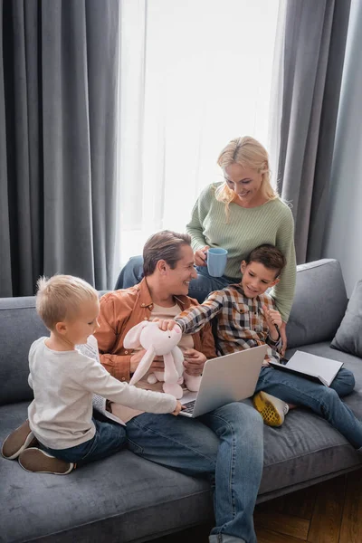 Glückliche Frau mit Söhnen hat Spaß in der Nähe von Mann auf Sofa mit Laptop sitzen — Stockfoto