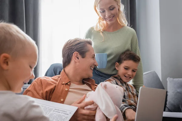 Donna sorridente con tazza di tè vicino al marito seduta sul divano con laptop, coniglietto giocattolo e bambini — Foto stock