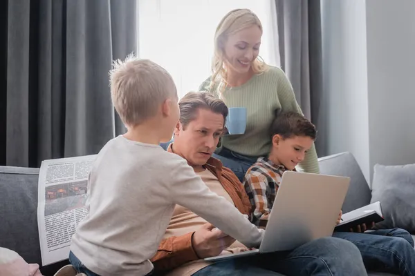 Man sitting on couch with laptop near kids with notebook and newspaper and woman standing with cup of tea — Stock Photo