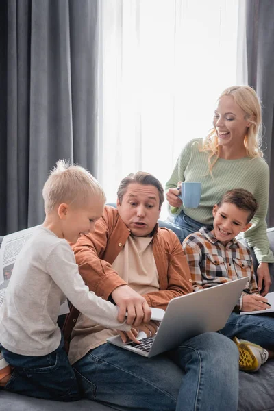 Hombre con el ordenador portátil y el teléfono inteligente divertirse con los niños cerca de esposa feliz con taza de té - foto de stock