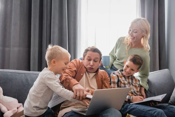 Fröhliche Kinder, die Spaß mit Papa haben, der auf dem Sofa mit Laptop neben fröhlicher Frau mit einer Tasse Tee sitzt — Stockfoto