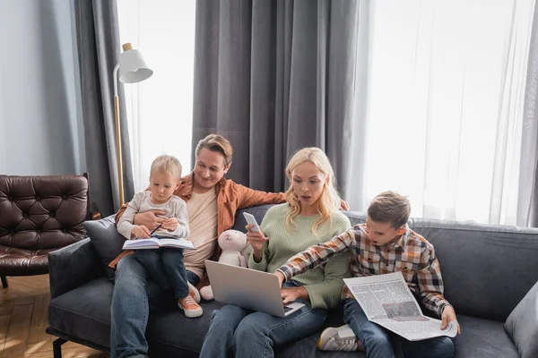 Ragazzo con giornale che punta al computer portatile vicino alla madre che lavora sul computer portatile sul divano — Foto stock