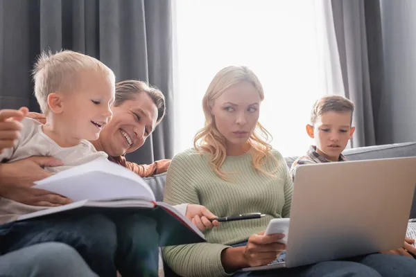 Happy man having fun with kids on sofa near displeased wife working on laptop — Stock Photo