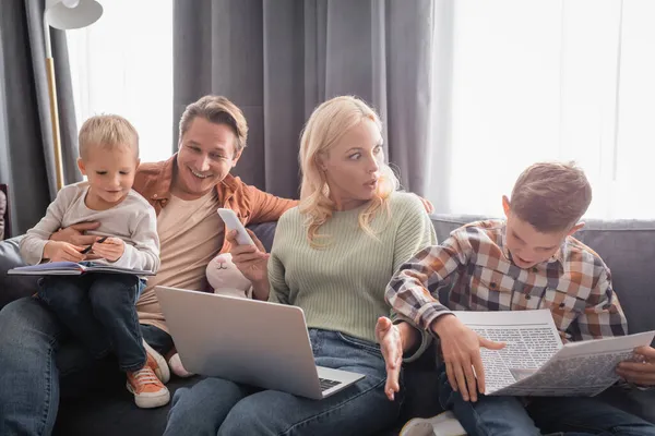 Шокована і зайнята жінка з ноутбуком і смартфоном, що працює біля веселої сім'ї, розважається на дивані — стокове фото