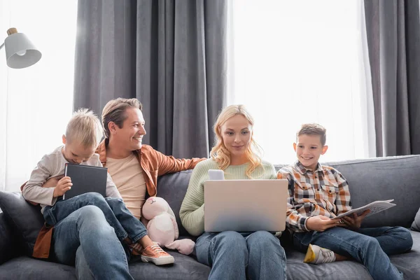 Frau mit Laptop arbeitet auf Sofa neben glücklichen Söhnen und Ehemann — Stockfoto