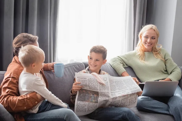 Alegre chico celebración periódico cerca de la familia en sofá en sala de estar - foto de stock