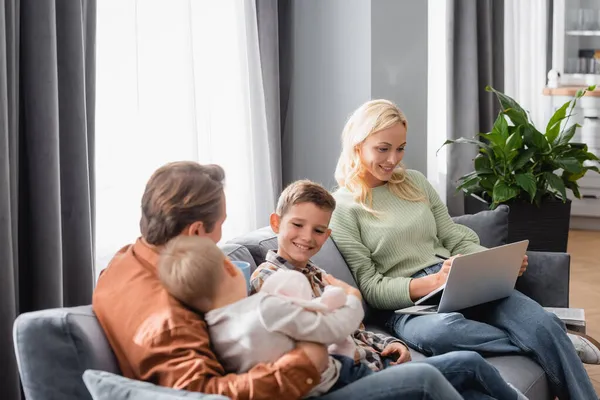 Улыбающаяся женщина с ноутбуком, работающая на диване рядом с веселыми сыновьями и мужем — стоковое фото