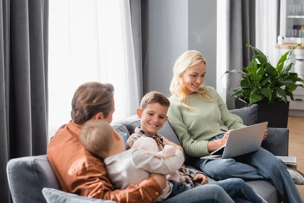 Smiling woman with laptop working on sofa near cheerful sons and husband — Stock Photo