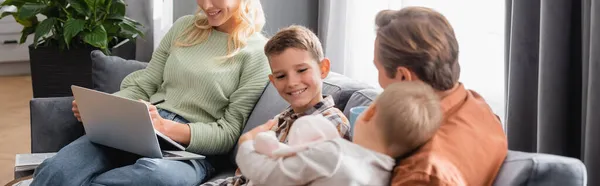 Feliz padre e hijos sentados en el sofá cerca de la mujer que trabaja en el ordenador portátil, pancarta - foto de stock